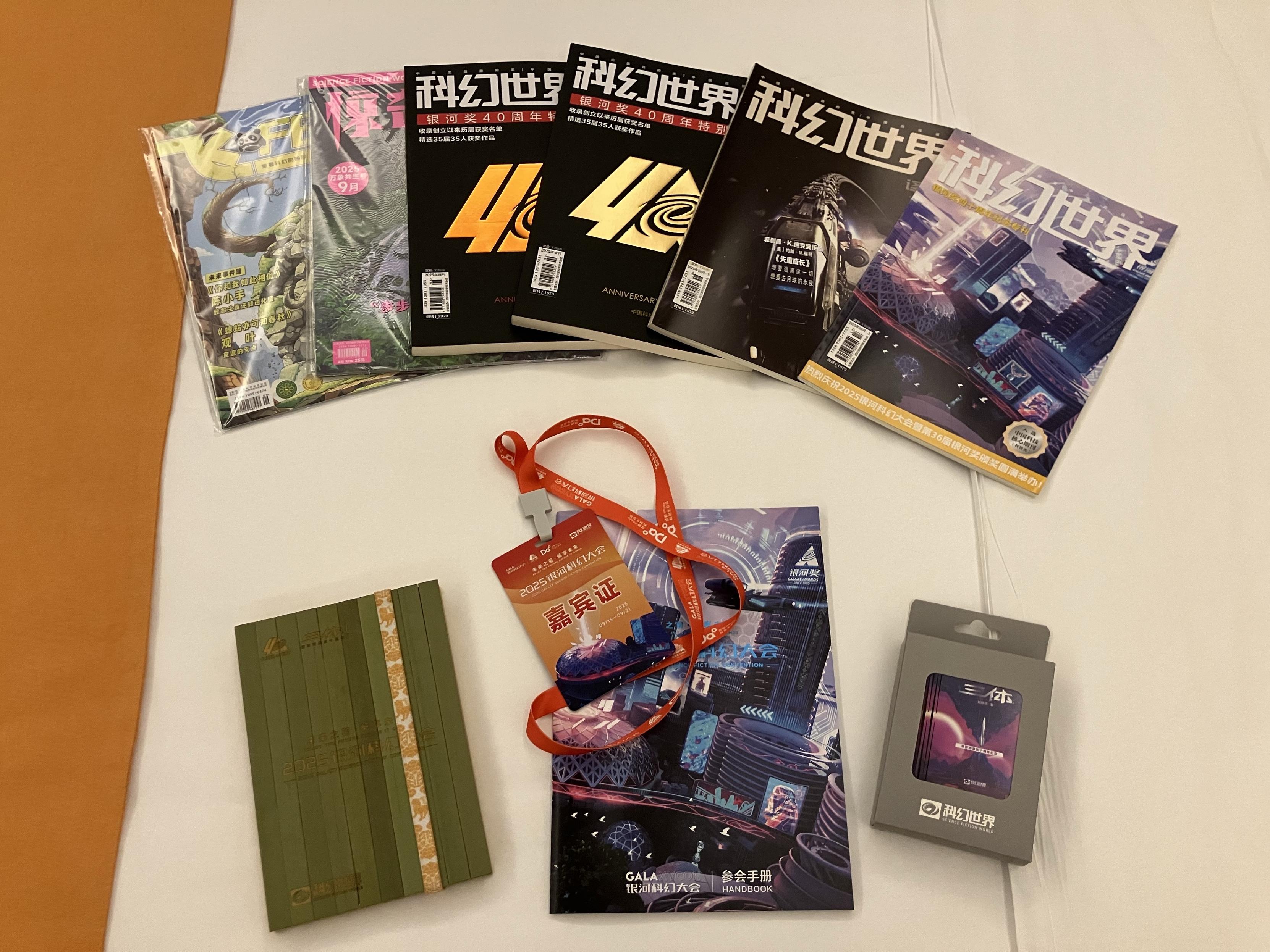 A collection of magazines, including special editions, laid out on a bed. Also present are a bamboo notebook, a badge with a red lanyard, and a small box, likely containing additional items or accessories. The background is a neutral surface.